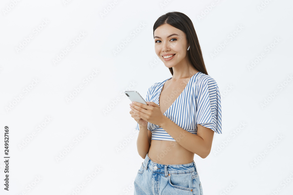 Indoor shot of sociable attractive happy asian female in cropped top holding smartphone wearing wireless earphones and smiling satisfied at camera listening music picking song in gadget over grey wall Indoor shot of sociable attractive happy asian female in cropped top holding smartphone wearing wireless earphones and smiling satisfied at camera listening music picking song in gadget over grey wall