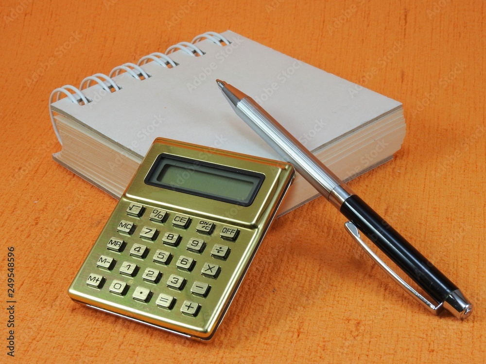 Close-up of a golden calculator, a small blank notepad and a pen on an orange surface. The notepad is closed. Close-up of a golden calculator, a small blank notepad and a pen on an orange surface. The notepad is closed.