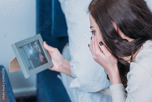 upset woman lying in bed, crying and looking at frame with picture of man at home