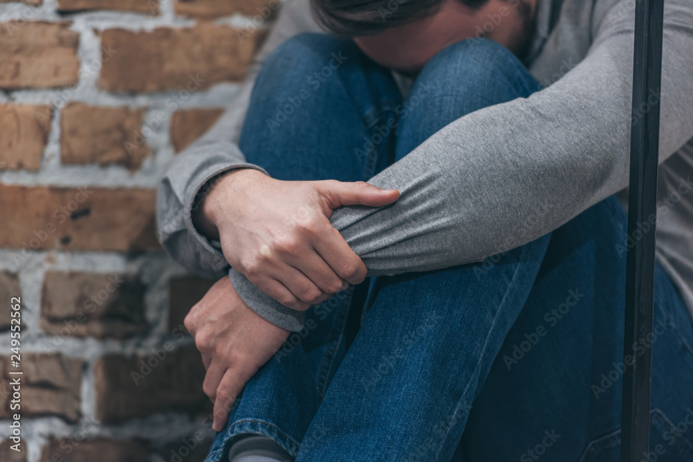 cropped view of man in gray sweater and blue pants sitting and hugging knees on brown textured background in room, grieving disorder concept cropped view of man in gray sweater and blue pants sitting and hugging knees on brown textured background in room, grieving disorder concept