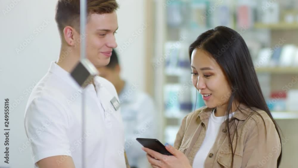Handheld medium shot of young Asian woman using smartphone at gadget store, shop assistant talking to her and helping her choose Handheld medium shot of young Asian woman using smartphone at gadget store, shop assistant talking to her and helping her choose
