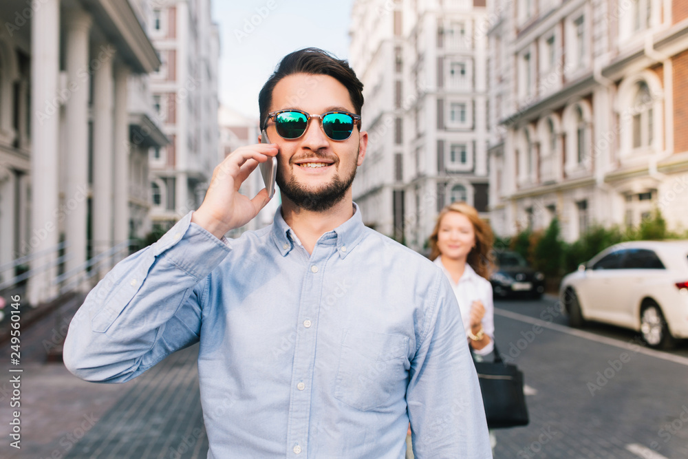Happy businessman in sunglasses speaking on phone on street. Pretty blonde girl catching him from behind on background Happy businessman in sunglasses speaking on phone on street. Pretty blonde girl catching him from behind on background