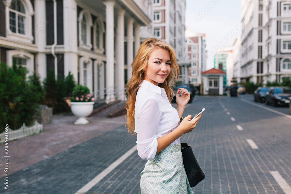 Pretty blonde business woman with long hair in white shirt walking on street around British quarter. She holds phone, smiling to camera