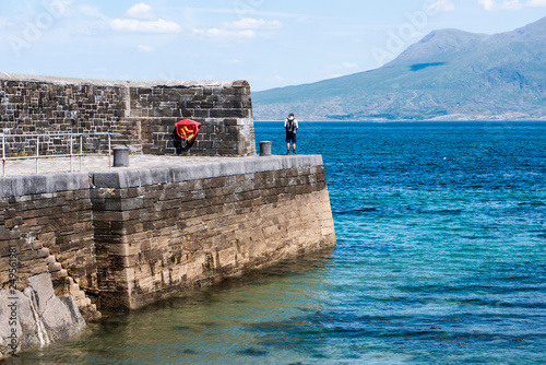 One man on an old stone pier on a sunny day, with turquoise oceans and mountains in the background. Taken in Renvyle along the Wild Atlantic Way in Ireland. photo