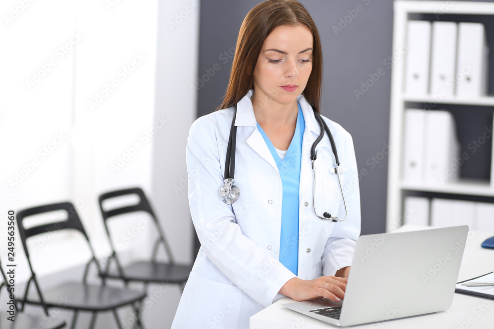Doctor woman at work. Portrait of female physician using laptop computer while standing near reception desk at clinic or emergency hospital. Medicine and healthcare concept Doctor woman at work. Portrait of female physician using laptop computer while standing near reception desk at clinic or emergency hospital. Medicine and healthcare concept