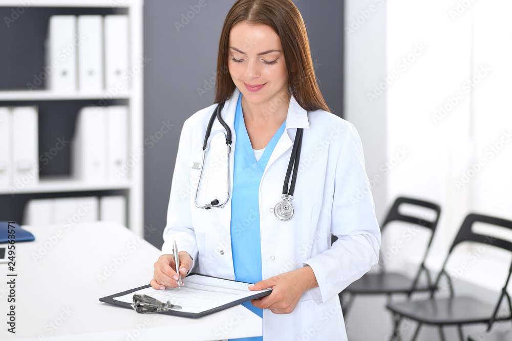 Doctor woman at work. Portrait of female physician filling up medical form while standing near reception desk at clinic or emergency hospital. Medicine and healthcare concept