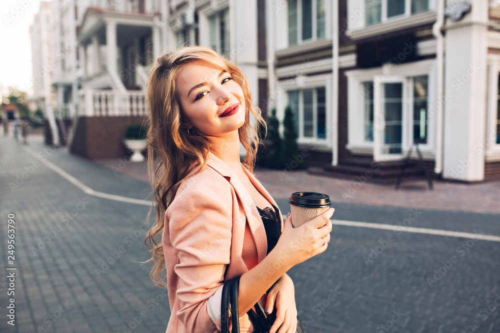 Fototapeta premium Closeup portrait attractive model with vinous lips walking with coffee in coral jacket on street. She smiling to camera