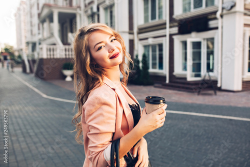 Closeup portrait attractive model with vinous lips walking with coffee in coral jacket on street. She smiling to camera