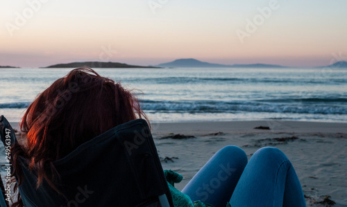 Woman sitting on a camping chair on a sandy beach watching a beautiful sunset over a calm ocean. Taken on Renvyle Beach along the Wild Atlantic Way in Ireland in summer. photo