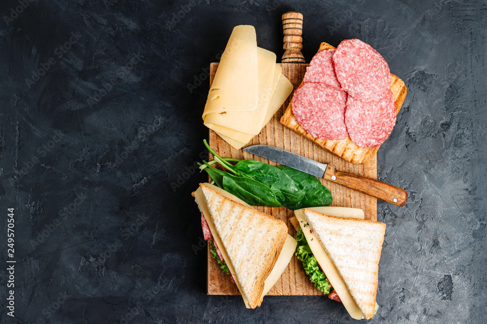 Sandwiches on a cooking board, grilled toast, salami sausage, salad lettuce and cheese on a dark background, top view