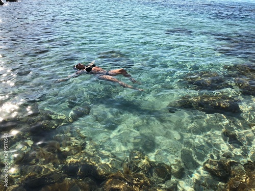 light-skinned girl in black swimsuit swims in the clear sea with colored stones and turquoise water, top view