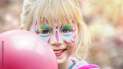 Glückliches geschminktes Mädchen beim Kinderkarneval auf einem Freizeitpark mit einem pinken Luftballon