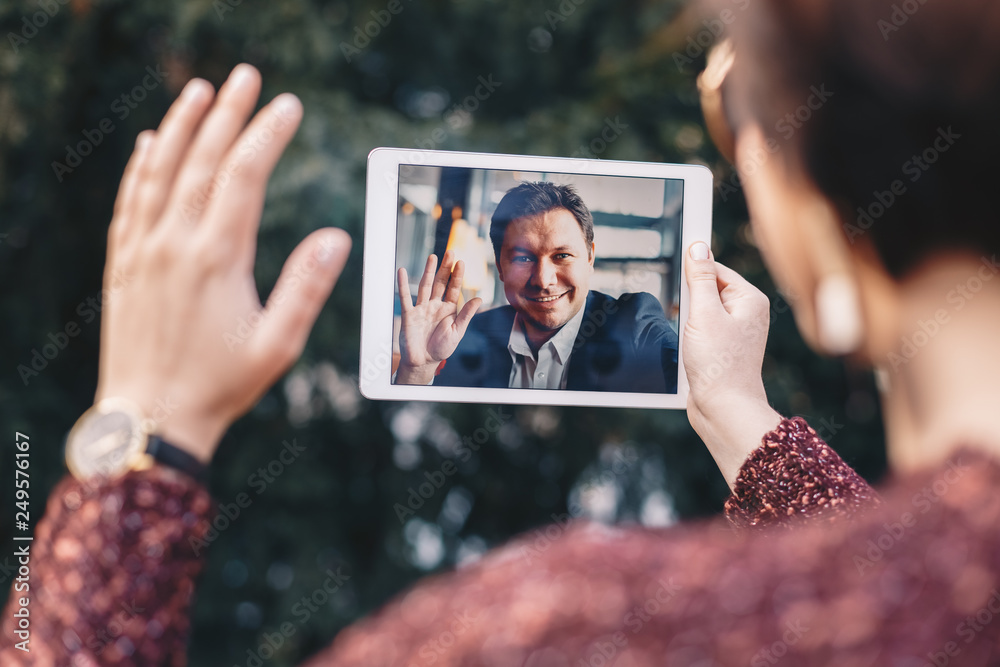 Young woman holding a tablet in her hands, having a video call chat with her freelancer boyfriend who is away on business trip. Concept of keeping long distant relationship in career oriented world.