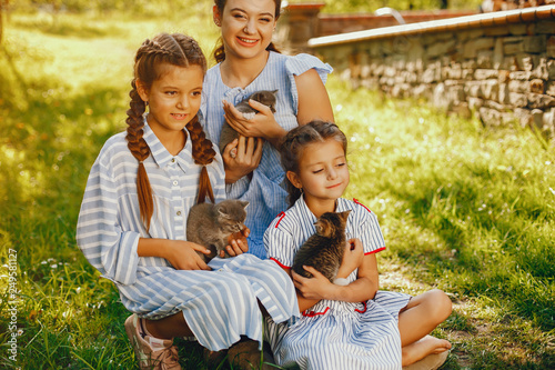 three beautiful and cute girls in blue dresses with beautiful hairstyles and make-up sitting in a sunny green garden and playing with a cats © prostooleh