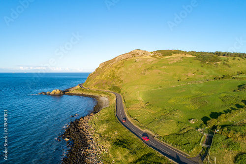 Northern Ireland, UK. Causeway Coastal Route a.k.a Antrim Coast Road near Ballygalley Head and resort with red cars. One of the most scenic coastal roads in Europe. Aerial view.