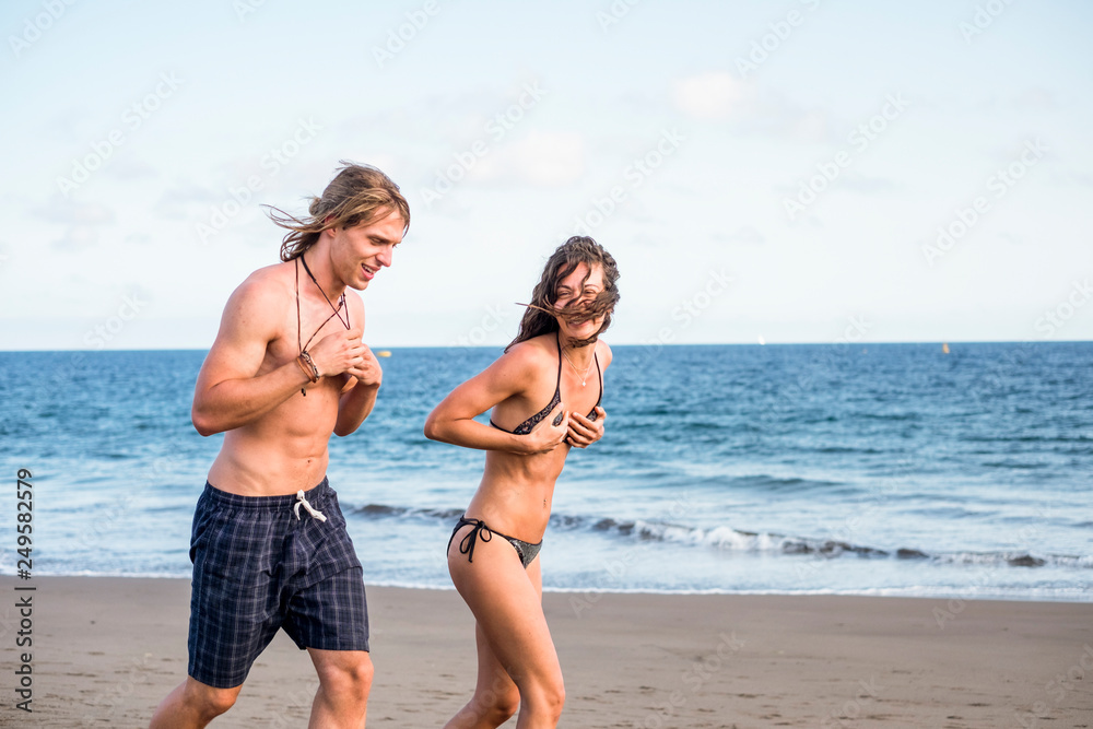 Beautiful young man and woman running for fun at the beach enjoying the summer vacation together - friendship and cheerful people laughing and enjoying the outdoor leisure activity- healthy lifestyle