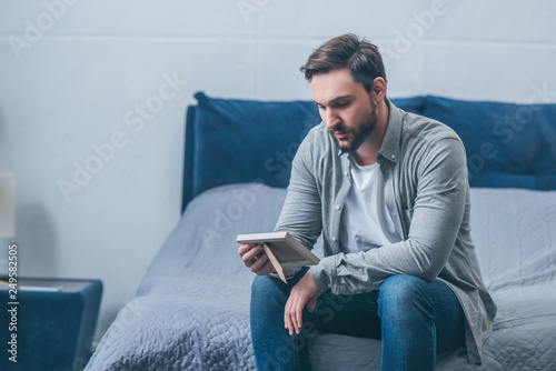 Obraz na plátně grieving man sitting on bed and looking at photo frame at home with copy space