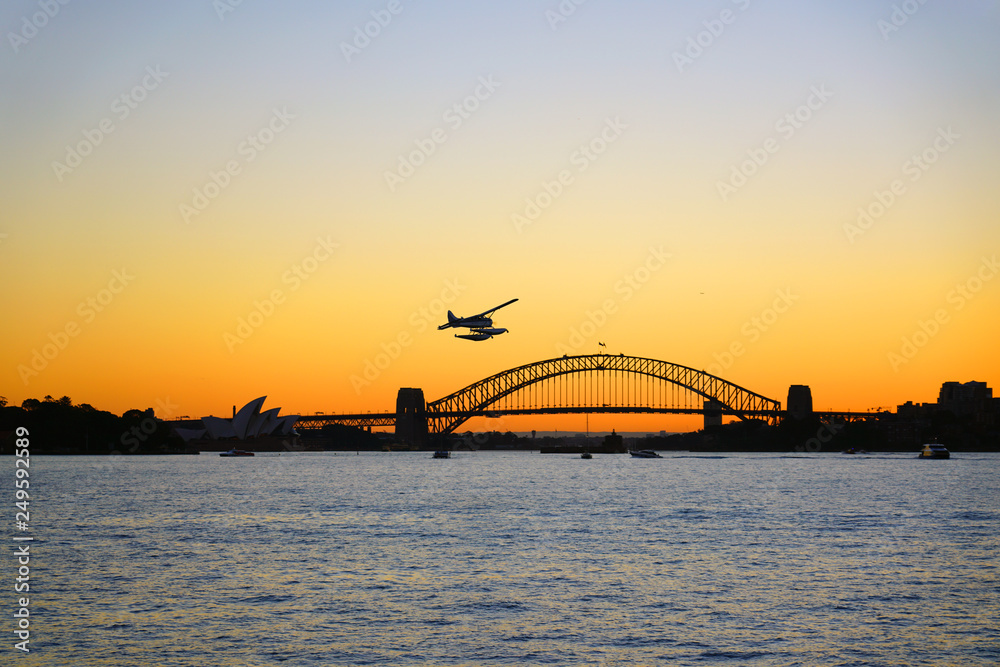 Naklejka premium Sunset view of a seaplane flying in the orange sky by the iconic steel Sydney Harbour Bridge in New South Wales, Australia