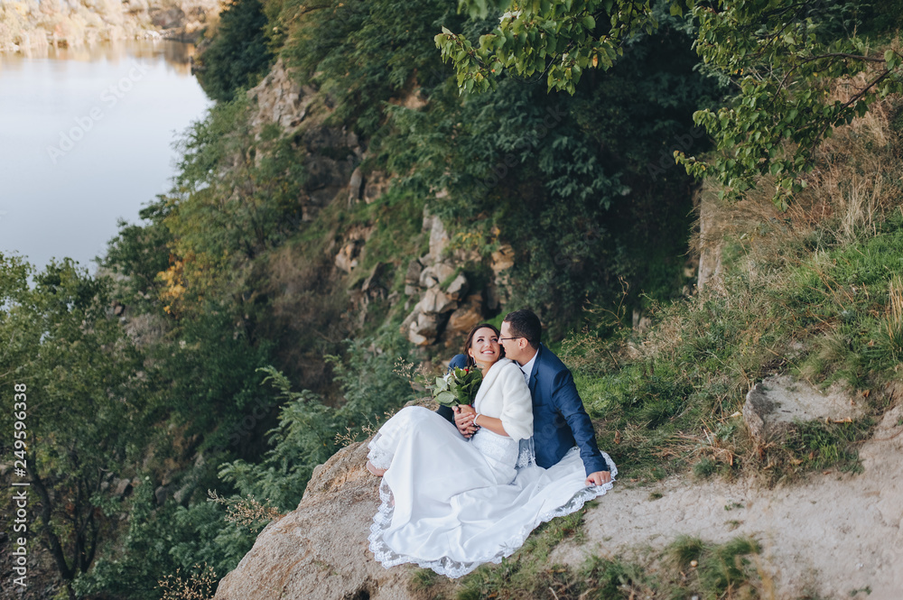 Beautiful newlyweds hugging on the background of rocks and a cliff. Portrait of a stylish groom with glasses and a cute bride in a white palette. Wedding photography.