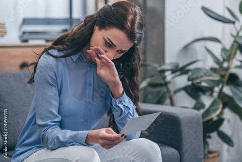 sad woman in blue blouse sitting on grey couch, crying and looking at photo in room, grieving disorder concept photo