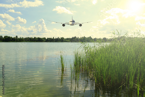 Canvas Print The plane flies in the blue sky with clouds over the lake and forest green sunset