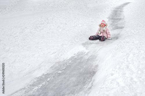 A happy little toothless 5-year old girl in warm clothes and woolen hat is going downhill from an ice hill in a winter park in the open air.