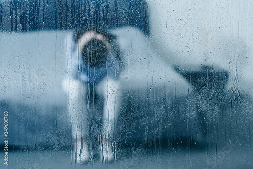 depressed woman sitting on bed and holding head in hands through window with raindrops