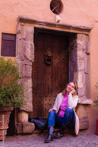 beautiful young girl is siting and talking on the phone near an ancient wooden door of a home / hotel in Rome, model with long blond hair, dressed in a white coat, pink sweater and jeans © Olga