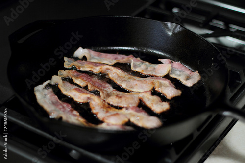 Strips of bacon frying in a cast iron pan on the stove.