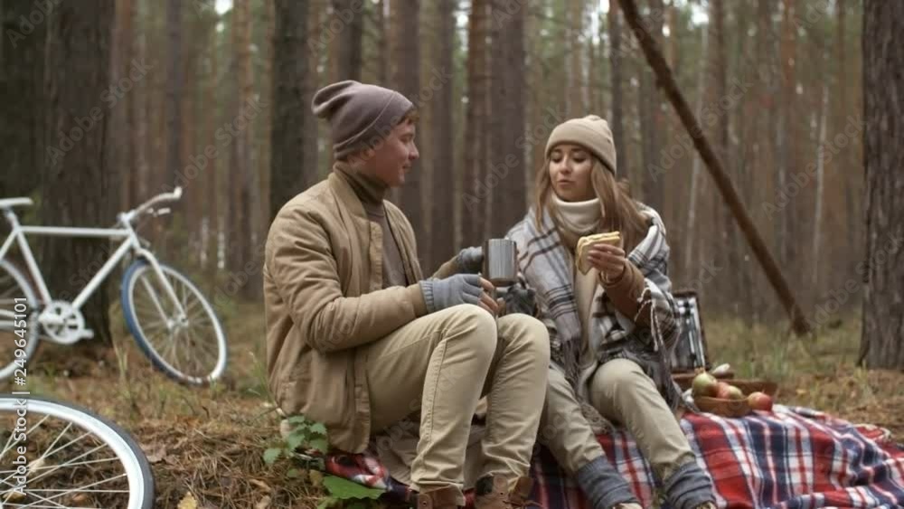 Zoom in of young couple in outerwear sitting on blanket in the forest and having picnic on autumn day: woman smiling and giving sandwich to boyfriend Zoom in of young couple in outerwear sitting on blanket in the forest and having picnic on autumn day: woman smiling and giving sandwich to boyfriend