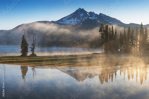 South Sister i Broken Top odbijają się nad spokojnymi wodami jeziora Sparks o wschodzie słońca w Cascades Range w środkowym Oregonie, USA, w świetle wczesnego poranka. Poranna mgła unosi się z jeziora w drzewa.
