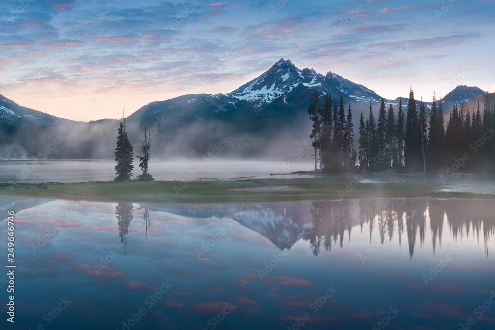 South Sister and Broken Top reflect over the calm waters of Sparks Lake at sunrise in the Cascades Range in Central Oregon, USA in an early morning light. Morning mist rises from lake into trees.  - obrazy, fototapety, plakaty 