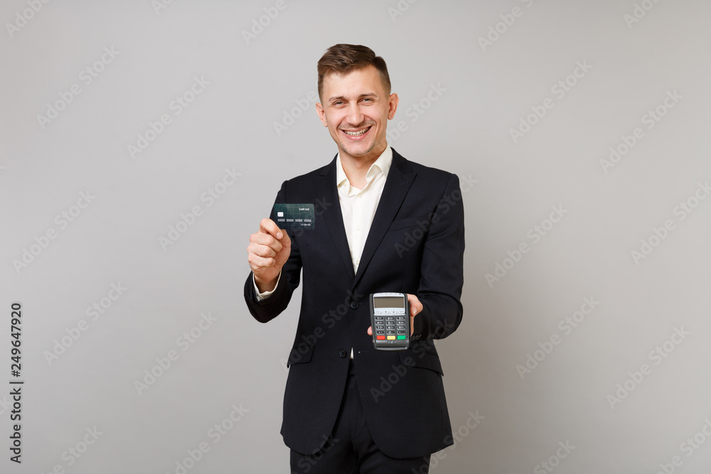 Joyful business man holding wireless modern bank payment terminal to process and acquire credit card payments, black card isolated on grey wall background. Achievement career wealth business concept.