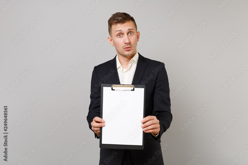 Bewildered young business man in classic black suit holding clipboard with blank empty sheet workspace isolated on grey wall background. Achievement career wealth business concept. Mock up copy space. Bewildered young business man in classic black suit holding clipboard with blank empty sheet workspace isolated on grey wall background. Achievement career wealth business concept. Mock up copy space.