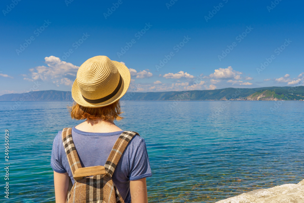 Tween tourist girl in hat and backpack admiring beautiful landscape of lake Baikal from waterfront of Listvyanka village in sunny summer day. Travelling, freedom and summer vacation concept