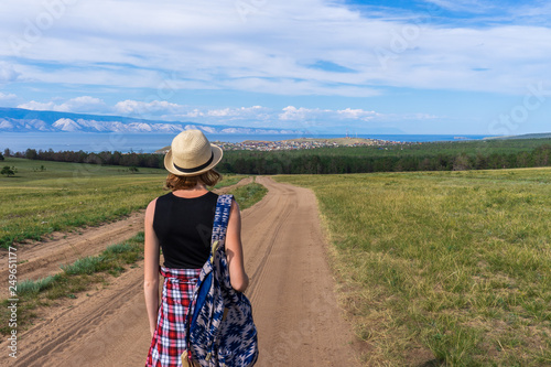 Tween tourist girl in hat and backpack walking on country road alone and admiring picturesque landscape of lake Baikal  mountains  village and blue sky. Travelling  hiking and summer vacation concept