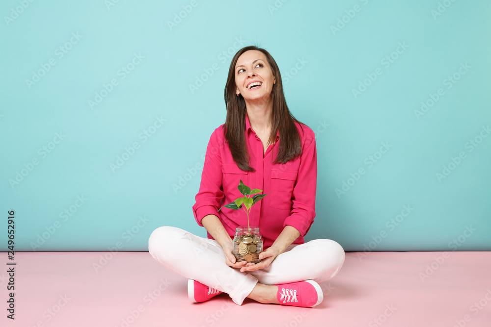 Full length of woman in rose shirt, white pants sitting on floor hold gold coins in glass jar isolated on bright pink blue pastel wall background studio. Fashion lifestyle concept. Mock up copy space. Full length of woman in rose shirt, white pants sitting on floor hold gold coins in glass jar isolated on bright pink blue pastel wall background studio. Fashion lifestyle concept. Mock up copy space.