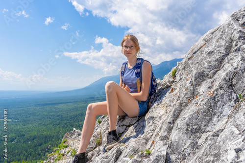 Tween tourist girl sitting on mountain cliff and looking at camera againt blue sky and summer landscape of valley. Hiking, travelling and wanderlust concept. Eastern Sayan, Buryatia, Russia