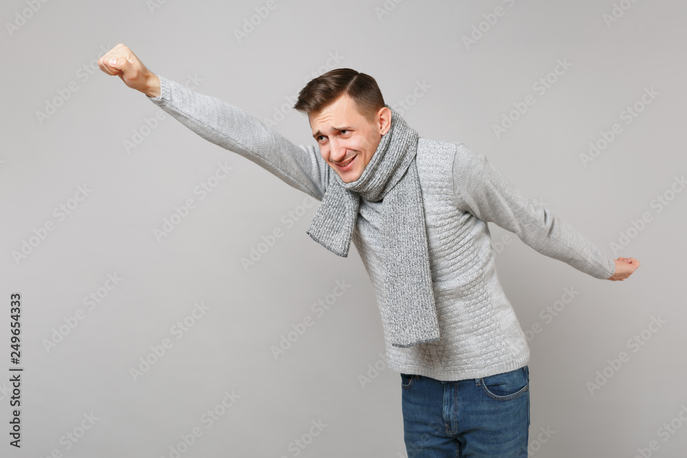 Funny young man in gray sweater, scarf with outstretched hand like Superman isolated on grey wall background. Healthy fashion lifestyle people sincere emotions cold season concept. Mock up copy space. Funny young man in gray sweater, scarf with outstretched hand like Superman isolated on grey wall background. Healthy fashion lifestyle people sincere emotions cold season concept. Mock up copy space.