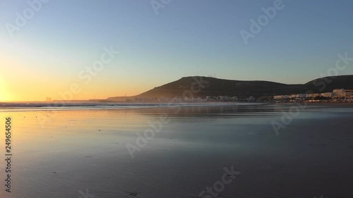Wide and flat Agadir Atlantic ocean beach at golden sunset, famous city landmark hill with inscription and old kasbah on the top seen in the background