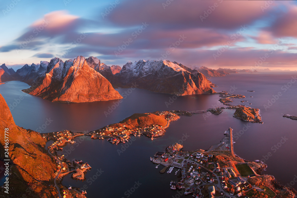 Winter landscape with houses in village, snowy mountains, sea, blue cloudy sky reflected in water at sunrise. Beautiful Hamnoy and Reine fisherman village, Lofoten islands, Norway. Norwegian fjords