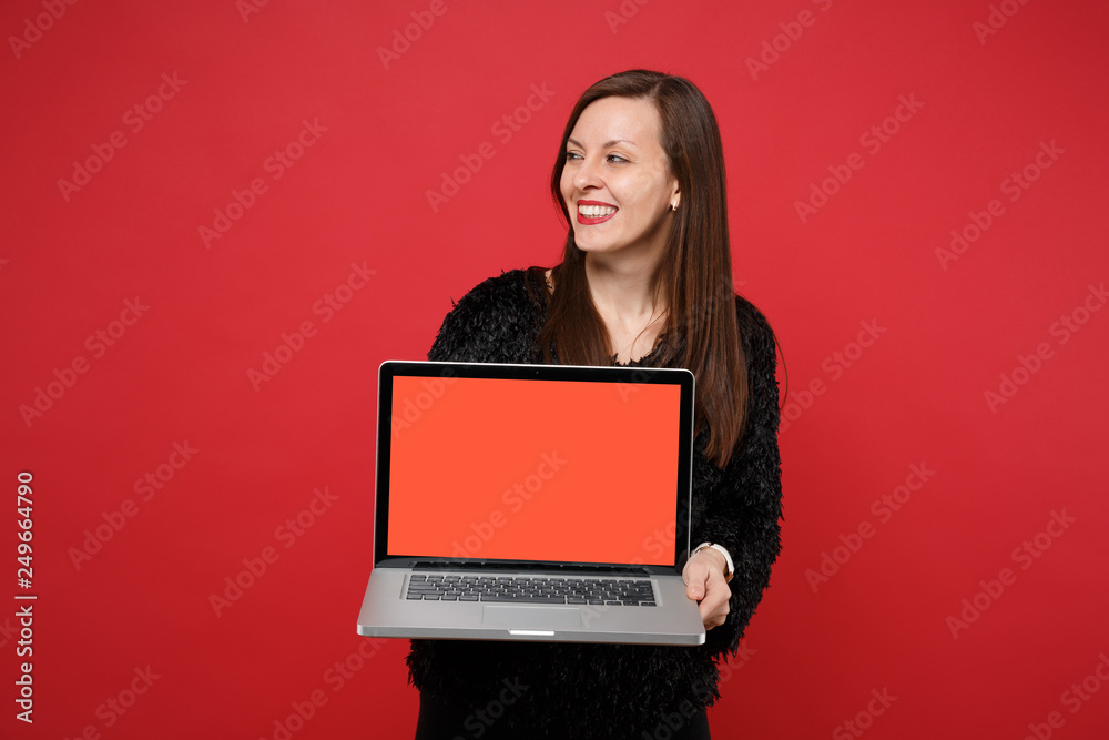 Naklejka premium Joyful young woman in black fur sweater looking aside hold laptop pc computer with blank empty screen isolated on bright red background. People sincere emotions, lifestyle concept. Mock up copy space.