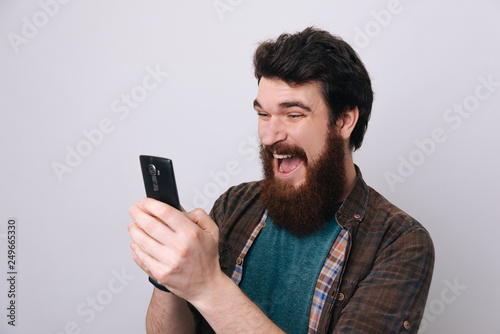 Young excited bearded man looking at his phone with surprise expression, isolated on gray background © Vulp
