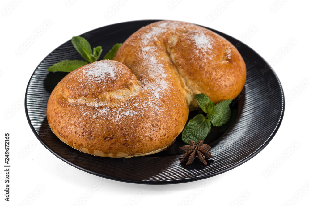 Fresh butter bun in the form of Latin letter S sprinkled with powdered sugar and decorated with fresh mint leaves, lying on a black plate isolated on a white background with shadows. Fresh butter bun in the form of Latin letter S sprinkled with powdered sugar and decorated with fresh mint leaves, lying on a black plate isolated on a white background with shadows.