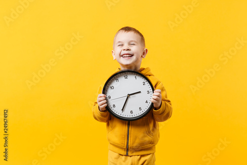 Little kid boy 3-4 years old wearing yellow clothes hold clock isolated on orange wall background, children studio portrait. People sincere emotions, childhood lifestyle concept. Mock up copy space.