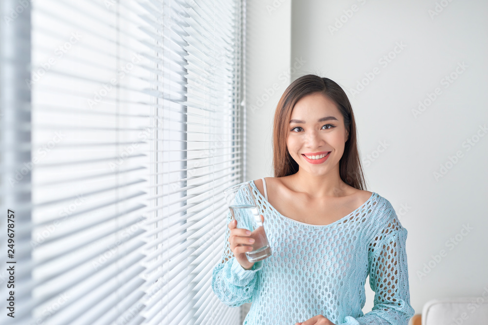 Young woman at home standing near window relaxing in her living room reading book and drinking water Young woman at home standing near window relaxing in her living room reading book and drinking water
