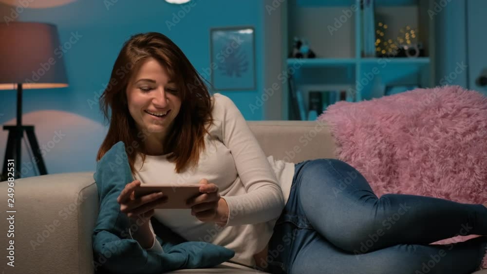 A pretty young girl using smartphone while lying on a couch in the living room at home in the night, flirting, texting while relaxing. Woman using an app on smartphone and reading news. A pretty young girl using smartphone while lying on a couch in the living room at home in the night, flirting, texting while relaxing. Woman using an app on smartphone and reading news.