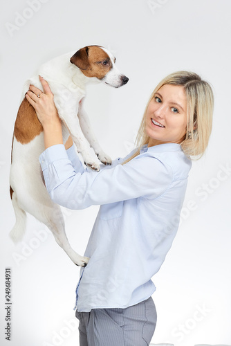 Blonde caucasian woman with smile watches straight holds in hands her Jack Russell Terrier on the white background.