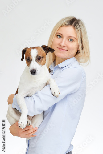 Blonde caucasian woman with smile watches straight hugs her Jack Russell Terrier on the white background.