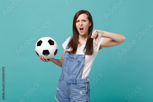 Irritated young woman football fan support favorite team with soccer ball showing thumb down isolated on blue turquoise background. People emotions, sport family leisure concept. Mock up copy space. © ViDi Studio